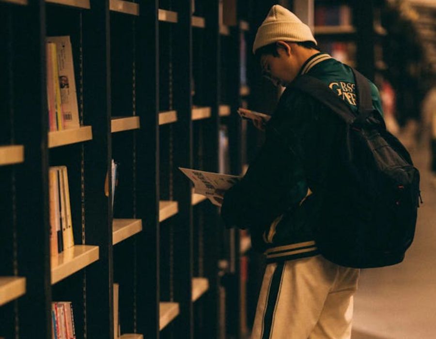 A boy in a Library Standing next to a Shelf and Holding a Book